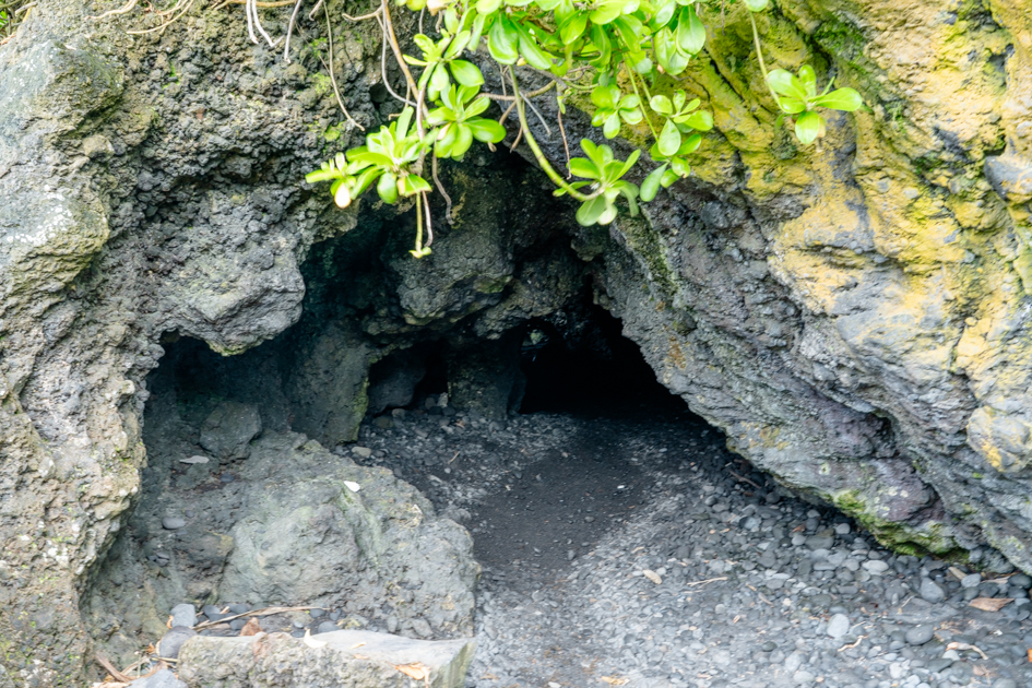 Entrance to a lava tube in Hawaii with a small crawl space opening and a distant view of the ocean through volcanic rock.