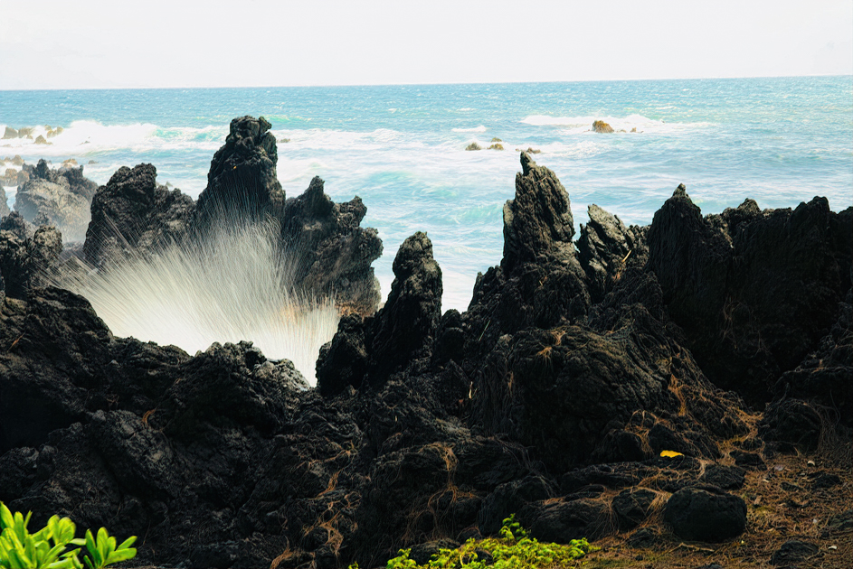 A powerful ocean blowhole shoots water high into the air through black lava rocks along the Hawaiian coastline, surrounded by crashing waves.