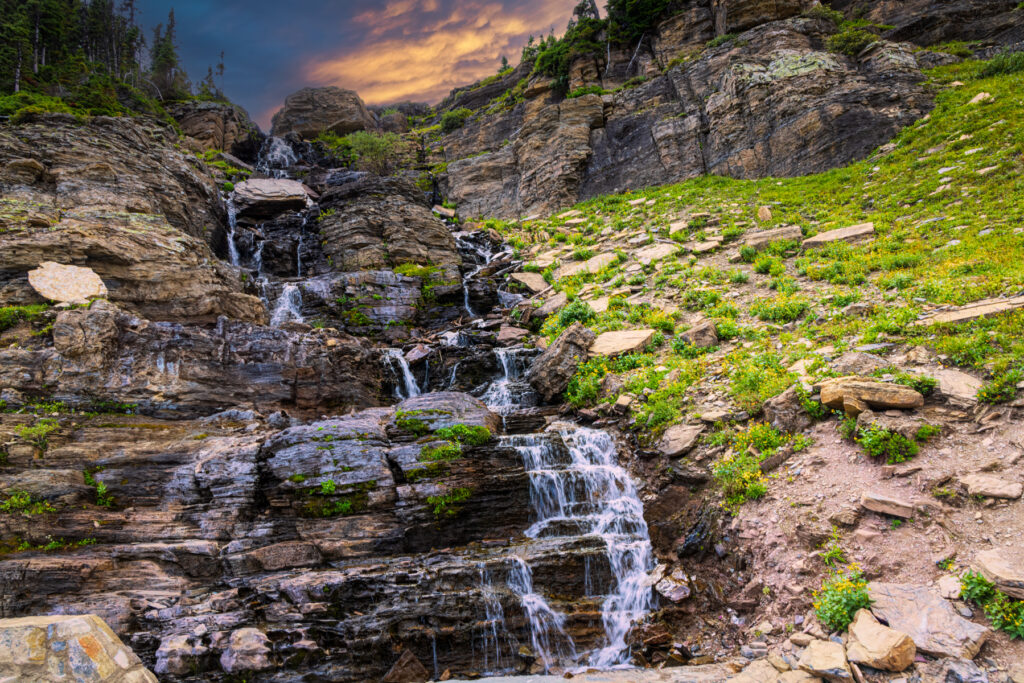 A waterfall cascading down rugged rocks along Going to the Sun Road in Glacier National Park, with the warm glow of sunset illuminating the scene.
