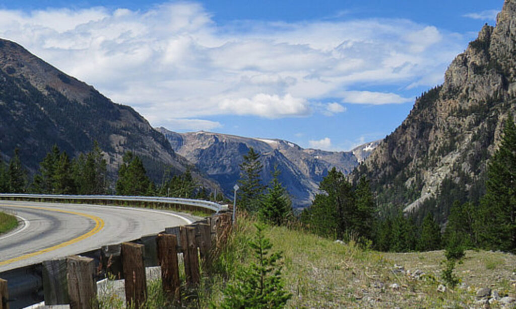 Curved mountain road on the Beartooth Highway with snow-capped peaks in the background under a clear blue sky.
