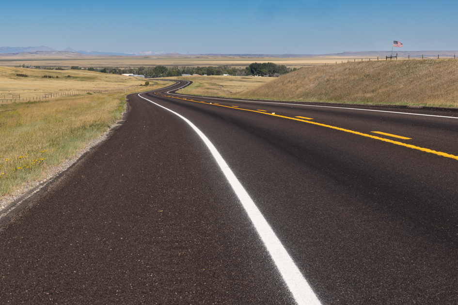 Long, empty road stretching through a desolate rural landscape under a wide open sky, photographed for Back Roads Lens travel blog