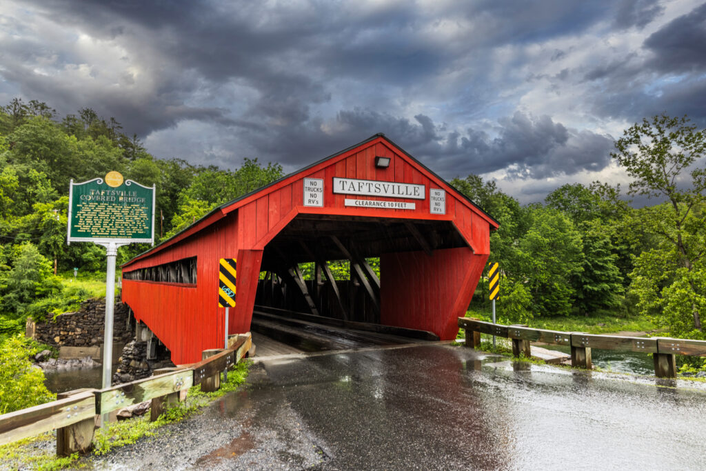 Taftsville Covered Bridge over the Ottauquechee River with red siding and Vermont countryside in summer light.  Northeast road trip