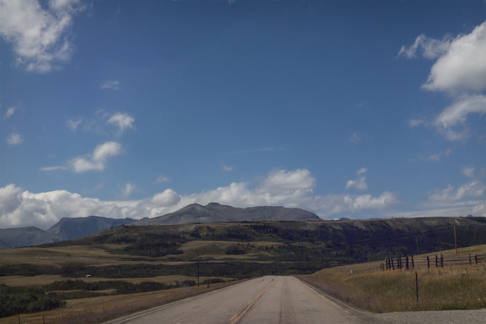 Empty rural road in Montana leading toward distant mountains under a vast sky, captured for Back Roads Lens photography blog