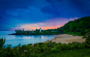 Sunrise over Waimea Bay in Oahu, Hawaii with orange and pink skies, a distant lighthouse, and fishermen on the beach.