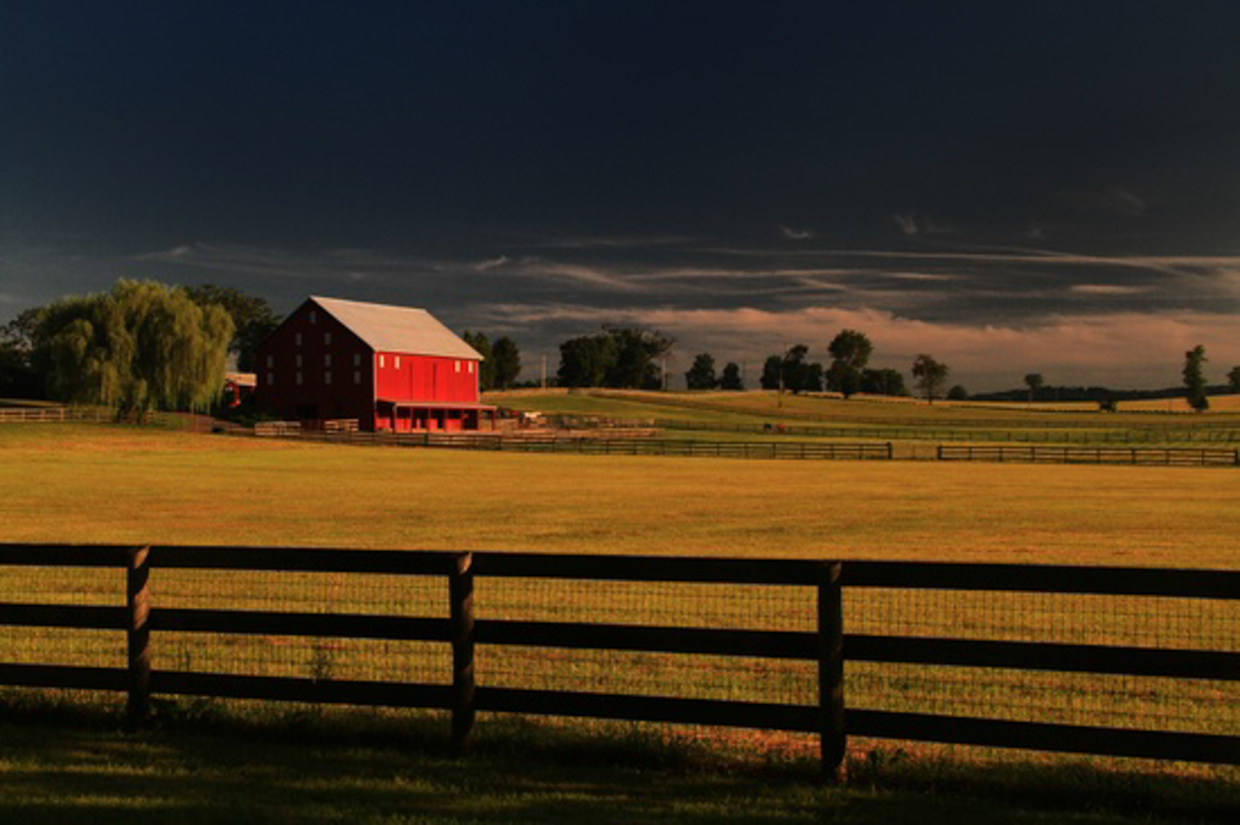 Red barn with a weathered wooden fence under a dark, stormy sky in a rural landscape.Back roads travel and photography blog. barn photography gallery