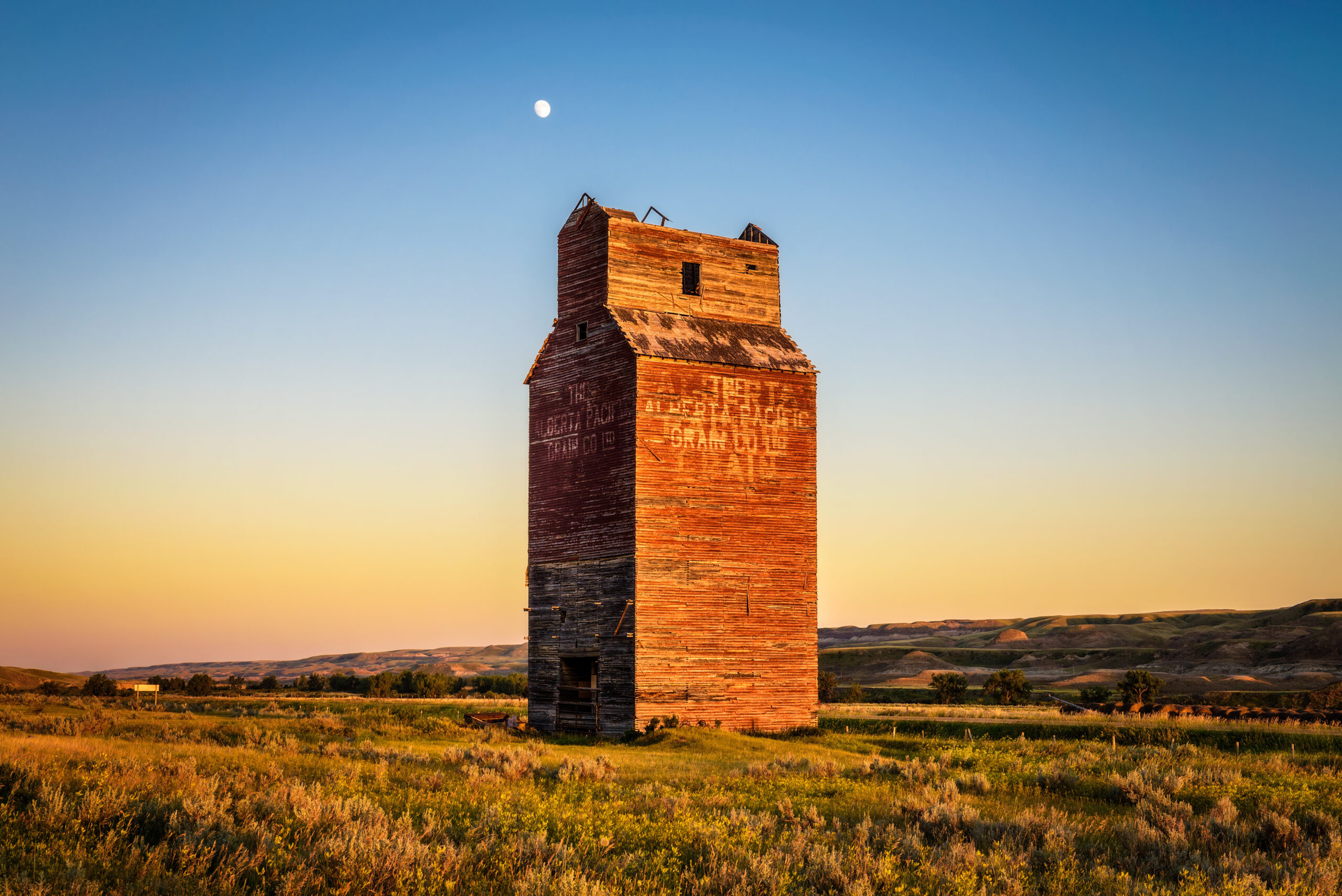 Weathered wooden barn with a tall, aged silo standing beside it in a rural landscape.