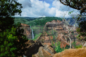 Colorful landscape of Waimea Canyon on Kauai with red cliffs, green vegetation, and a waterfall flowing into the valley.