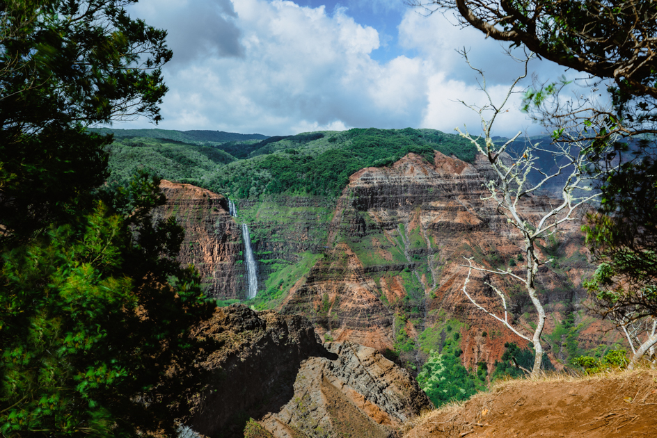 Colorful landscape of Waimea Canyon on Kauai with red cliffs, green vegetation, and a waterfall flowing into the valley.