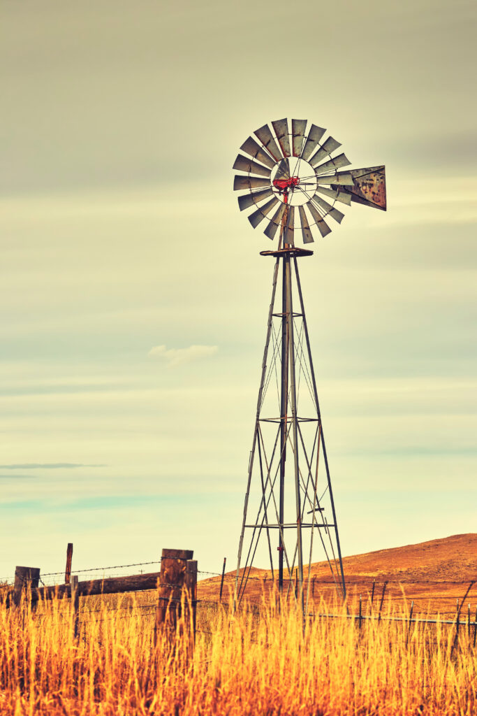 Close-up of a weathered windmill with a rusted metal fence in the foreground, set against Montana's expansive prairie landscape