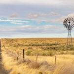 Old wooden windmill standing beside open fields with a quiet country road stretching into the distance.