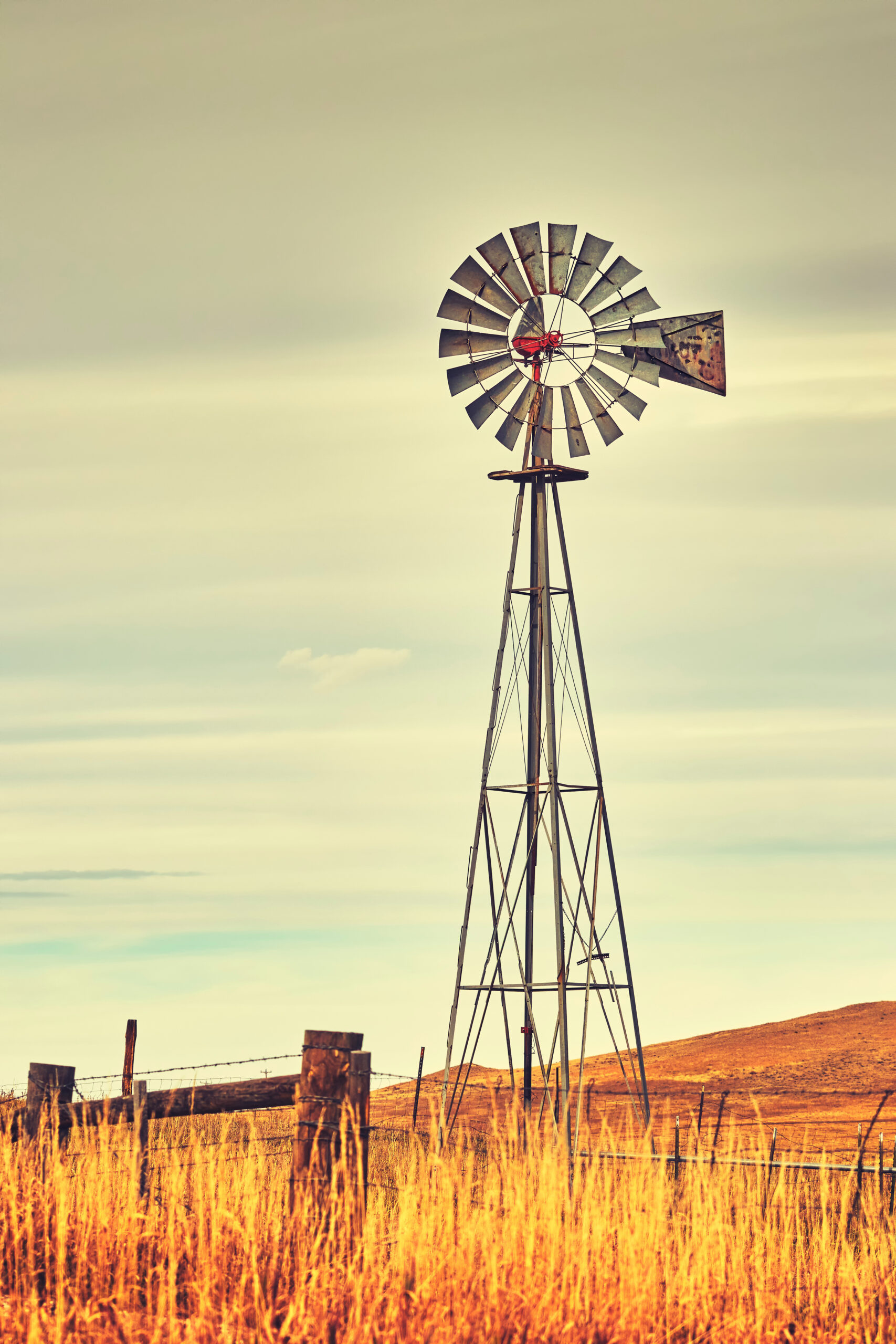 Close-up of a weathered windmill with a rusted metal fence in the foreground, set against Montana's expansive prairie landscape