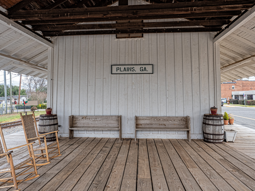 Two wooden rocking chairs on the porch of the historic train depot in Plains, Georgia, with “Plains, GA” signage in view.
