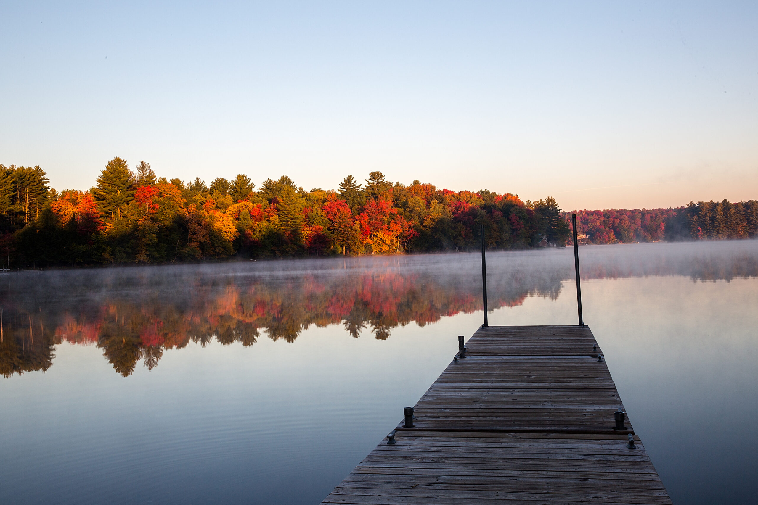 Wooden dock extending into White Lake at sunrise with fall foliage and reflections in the calm water