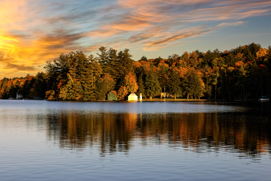 "Yellow boathouse reflected in a calm lake at sunset, surrounded by vibrant fall trees."