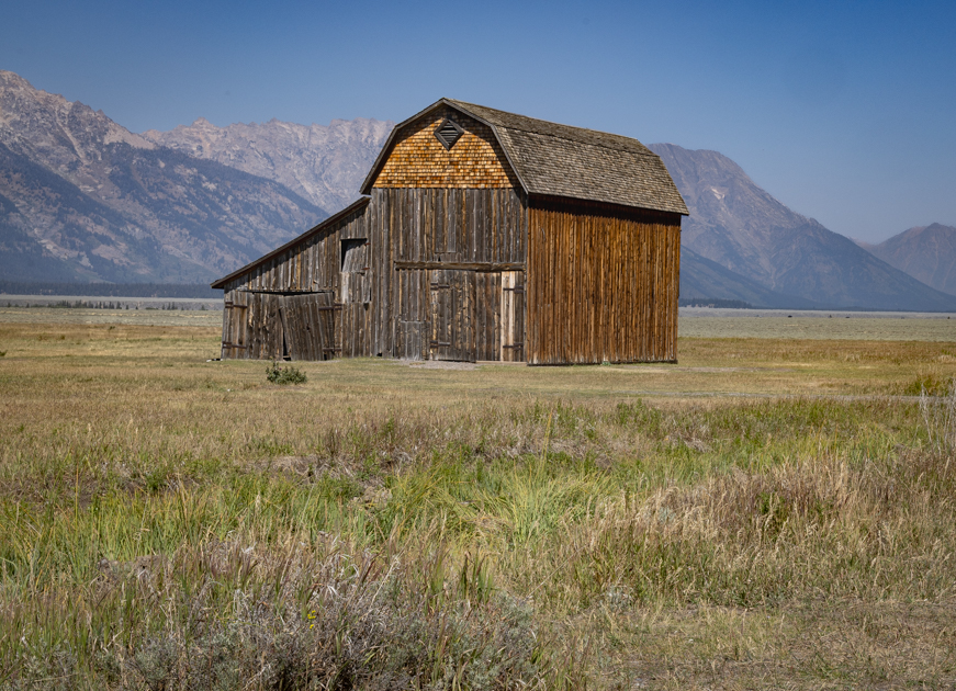 The rustic Thomas Murphy Barn on Mormon Row framed by sagebrush and distant peaks of the Teton Range under a bright blue sky
