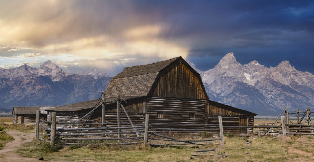 A dusty gravel road leads toward the historic John Moulton Barn with the snow-capped Teton Range rising behind it in Grand Teton National Park
