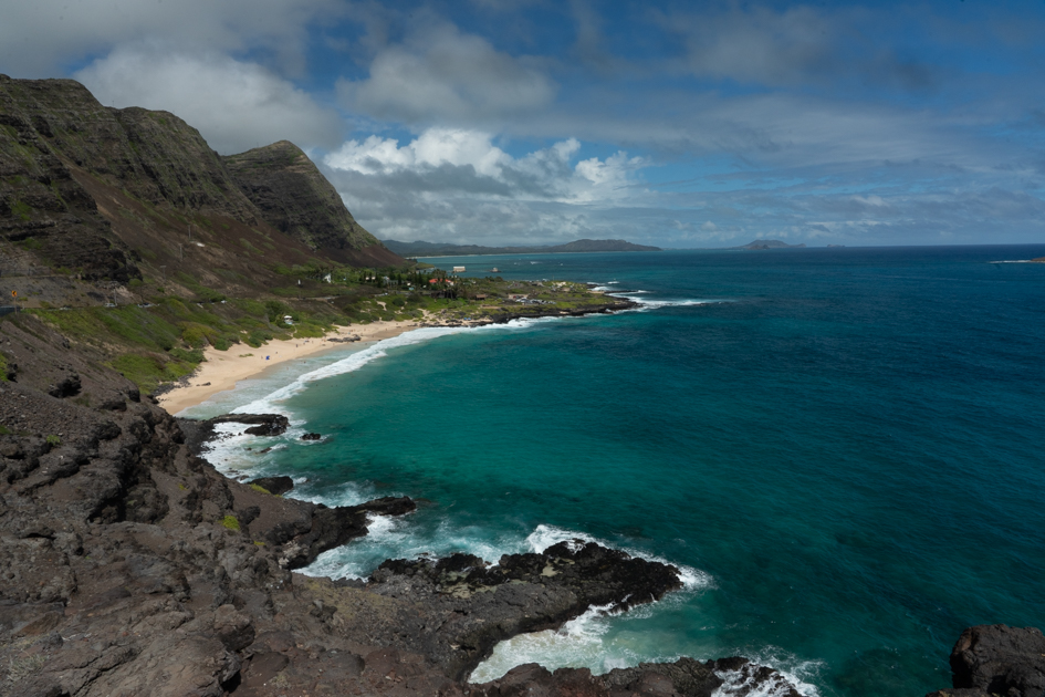 This photograph captures the historic Makapuʻu Point Lighthouse, built in 1909, as seen from the trail above the cliffs on Oahu’s southeastern coast. The bright red roof contrasts against the deep blue Pacific, with rugged lava rock and distant islands visible in the background. A classic stop for hikers and photographers exploring the island’s wild shoreline.