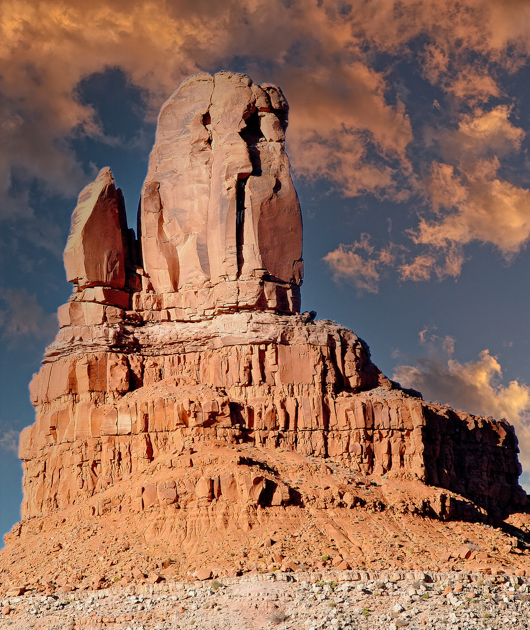 Owl Rock rising from the red desert floor in Monument Valley, framed by deep blue sky and scattered desert brush.