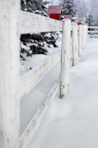 Red birdhouse perched on a white picket fence post surrounded by fresh snow.