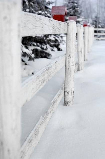 Red birdhouse perched on a white picket fence post surrounded by fresh snow.